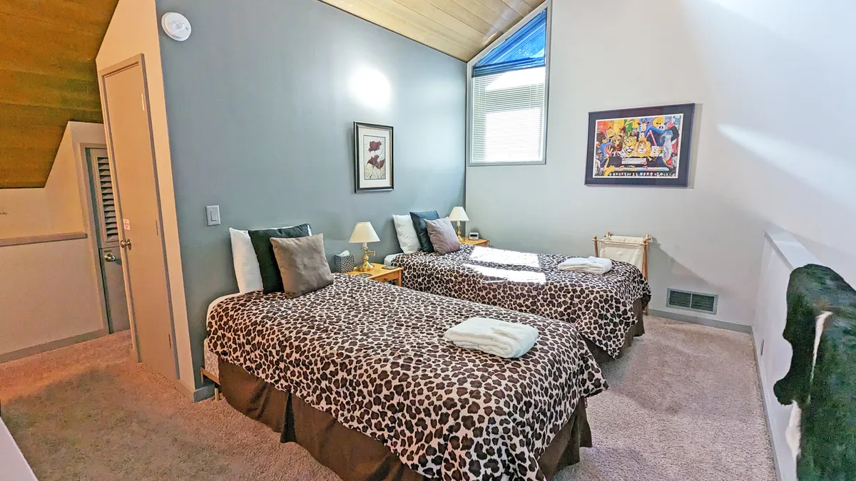 An airy loft space with two twin beds covered in playful leopard print bedding, cheerful artwork on the wall, and a window letting in plenty of natural light. The arrangement is perfect for kids or teens, offering a fun and welcoming vibe.