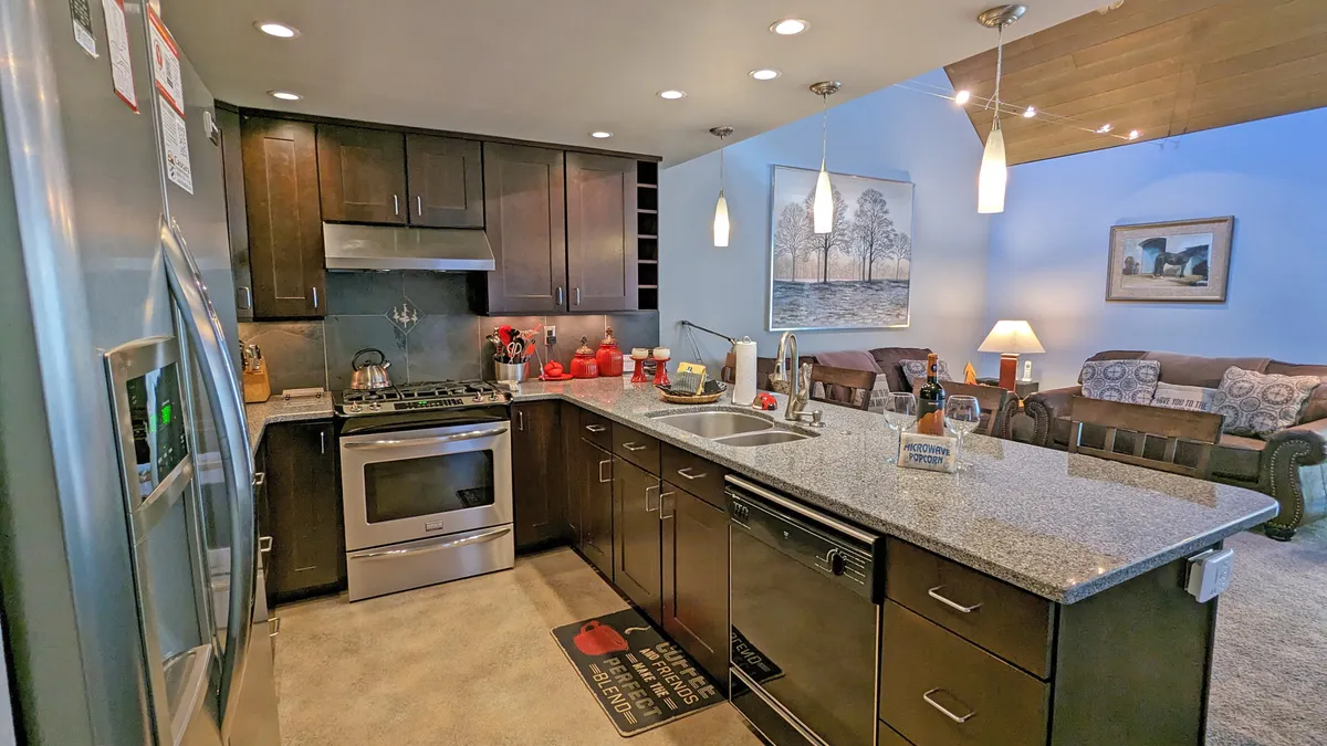 An inviting open-plan kitchen with granite countertops, stainless steel appliances, and red kitchenware visible on the counters. The design allows views into the attached living area, suggesting an easy transition from indoor to outdoor activities and providing a comfortable space for social interaction.