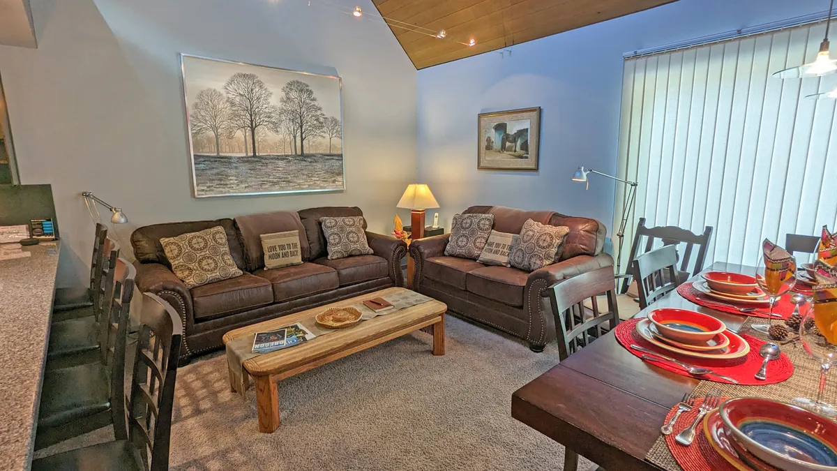 An expansive open-concept living and dining area with leather sofas, a wooden coffee table, a dining table set for six, and tasteful artwork. The space is warm and sunlit, with comfortable seating clustered around a gas fireplace, ideal for relaxation and conversation after a day of adventure.