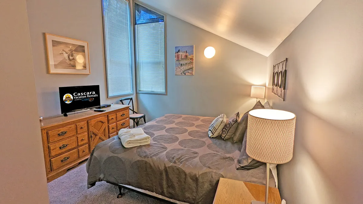A bright, welcoming bedroom featuring a bed adorned with decorative pillows, a cozy nightstand with a lamp, a television for entertainment, and a dresser topped with artwork. Sunlight filters softly through the window, creating a restful ambiance.