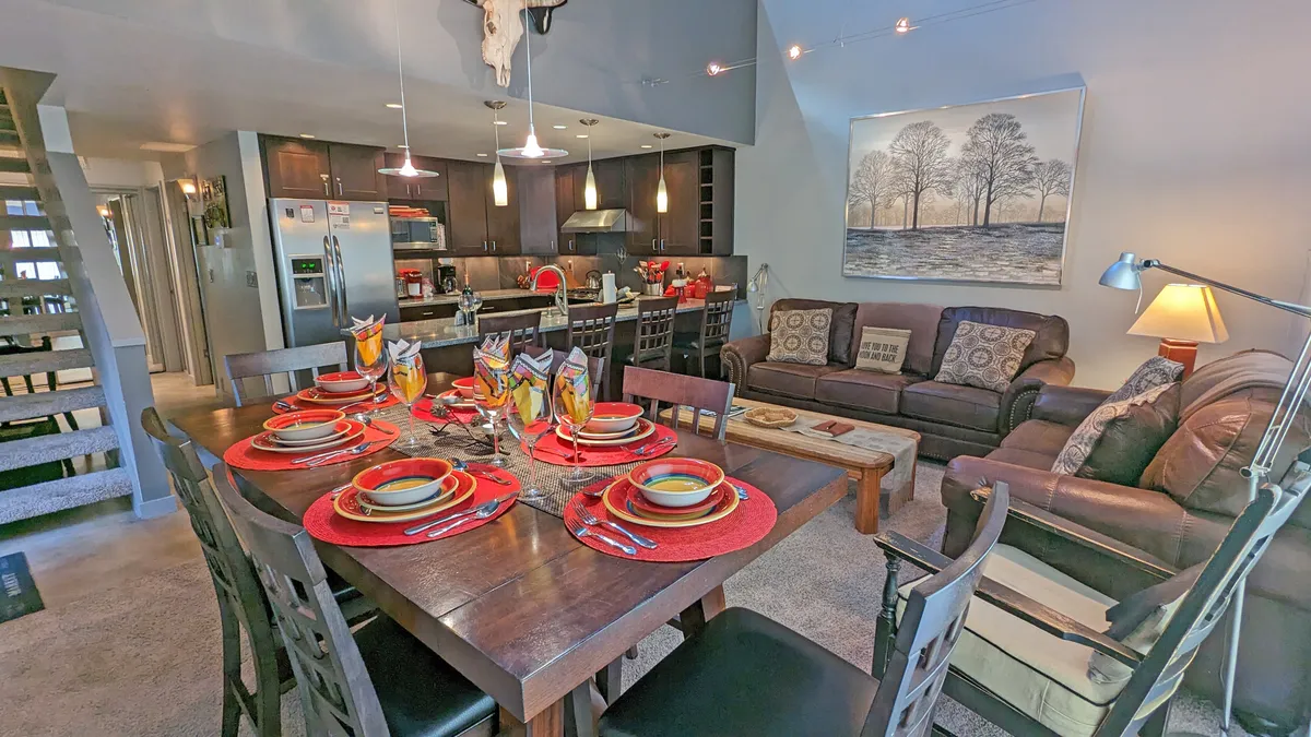 The image showcases a warm, sunlit open-concept living space featuring a colorful dining table set for a meal, a comfortable seating area perfect for relaxing or game nights, and a modern kitchen island ideal for cooking together. The ambiance is inviting, ideal for group gatherings and cozy evenings after outdoor adventures.