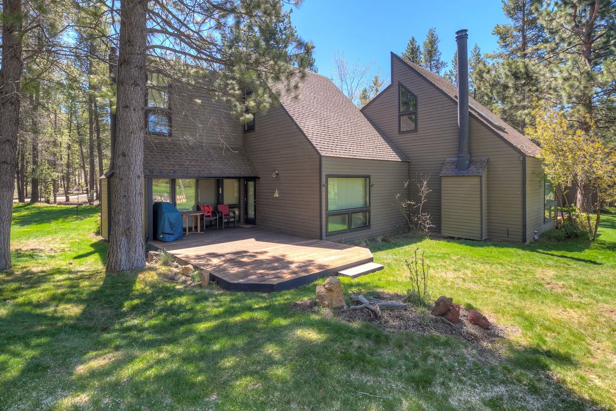 A tranquil, expansive wooden deck stretches out from the house, bordered by large windows and surrounded by towering pine trees. The deck is designed for outdoor dining and relaxation, with views ideal for sunrise coffees, sunset gatherings, and stargazing under clear Central Oregon skies. The natural scenery creates a peaceful, immersive environment.