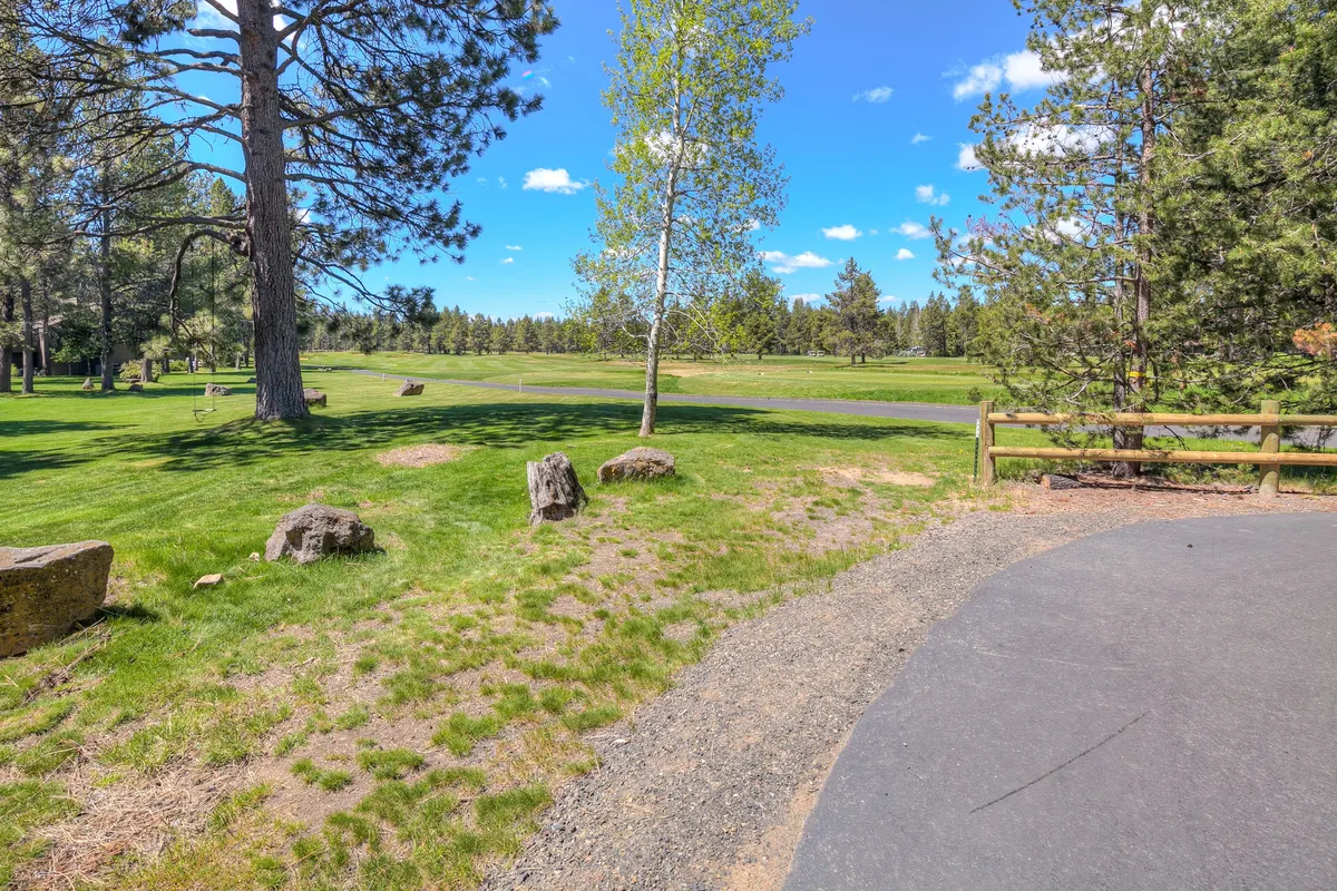 A serene exterior scene with tall trees, lush green grass, rock formations, and a gently curving pathway. The area feels open, peaceful, and perfect for outdoor activities like walking, picnicking, or golfing.
